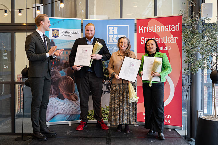 Fyra personer p&aring; en scen vara tre h&aring;ller i diplom och blommor.