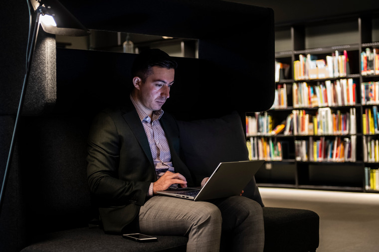 Manlig student sitter med dator i en soffa i biblioteket.