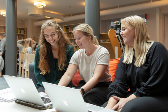 Tre tjejer sitter i grå soffa i biblioteket.