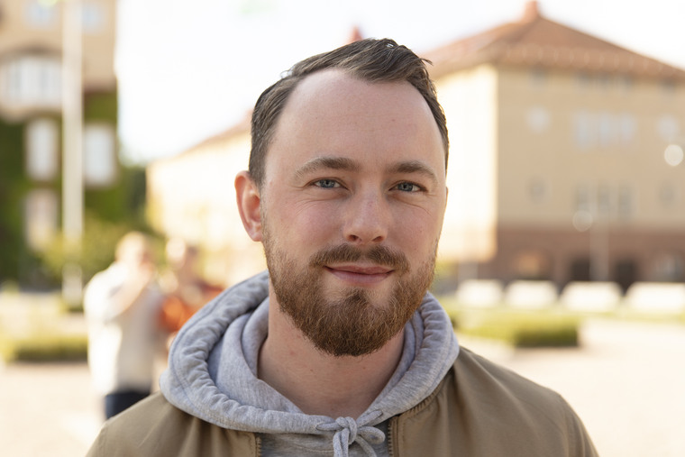 Kille med mörkt hår och skägg står med hus 4 i bakgrunden.