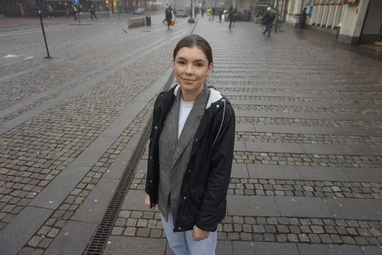 Emelie tittar leendes in i kameran, ståendes på en gågata en grå vinterdag.