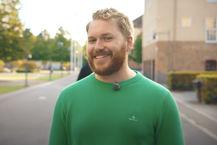 Leende kille i gr&ouml;n tr&ouml;ja st&aring;r utomhus p&aring; h&ouml;gskolans campus.