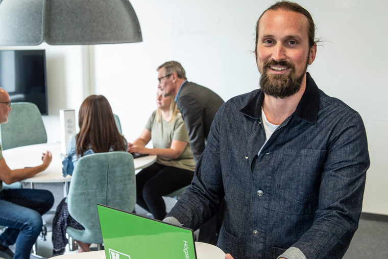 En person i förgrunden med dator, och personer runt ett bord i bakgrunden.