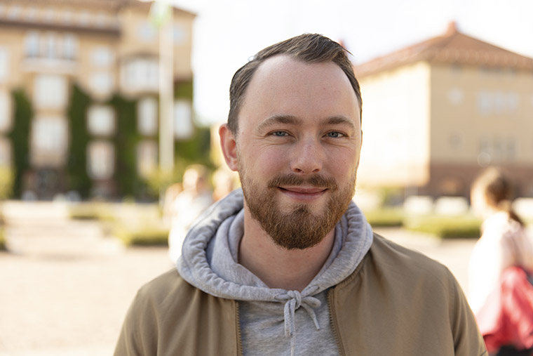 Kille utomhus på högskolans campus, tittar leende in i kameran.