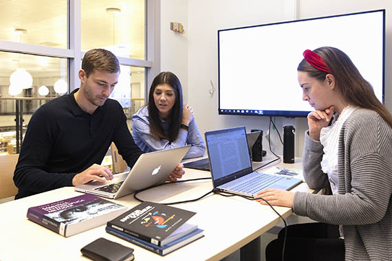 Tre studenter grupparbetar med böcker och datorer